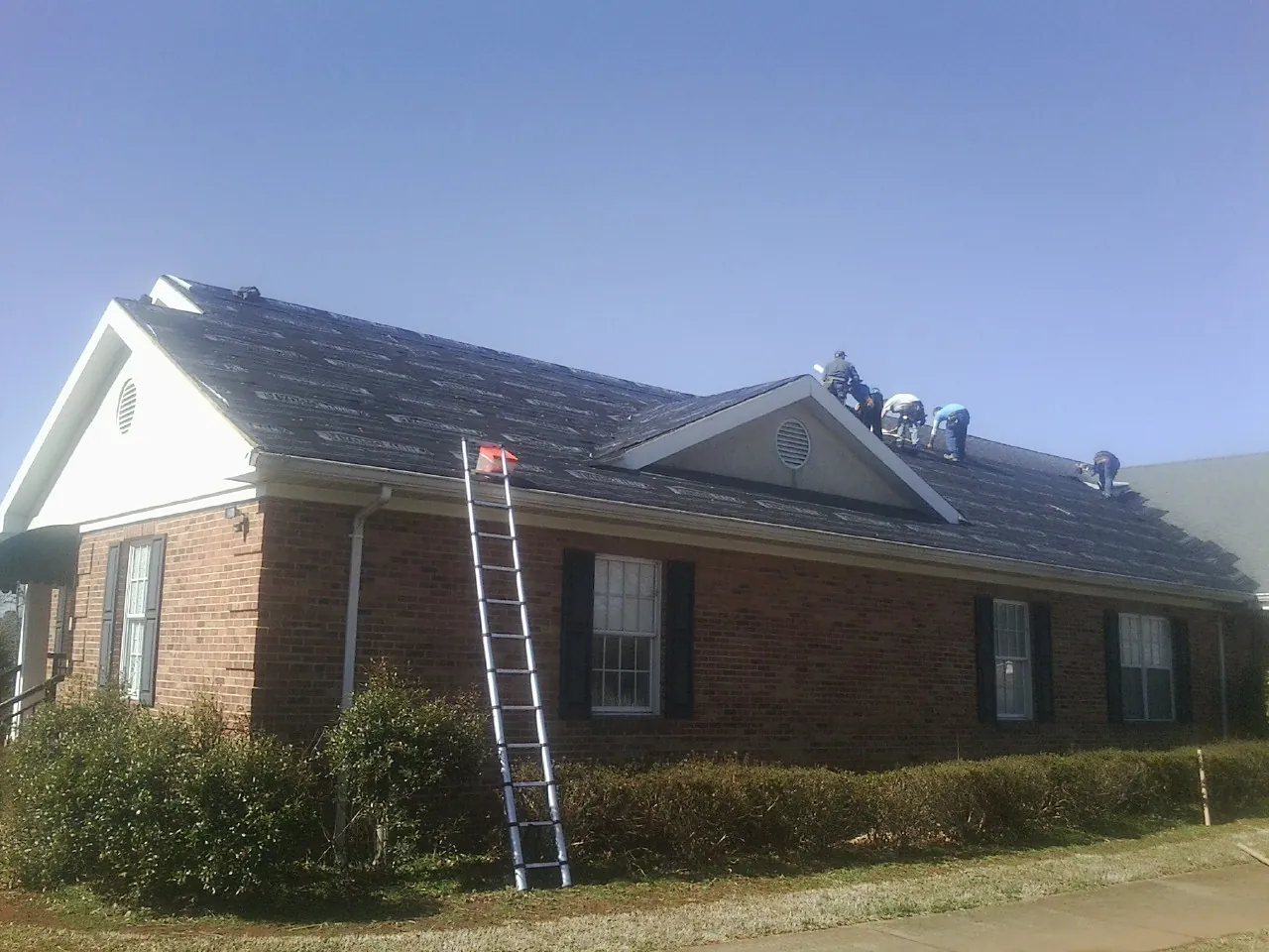 Crew tearing shingles off a brick ranch roof with a ladder set — Metal Roof Repair work by our crew in Delmont